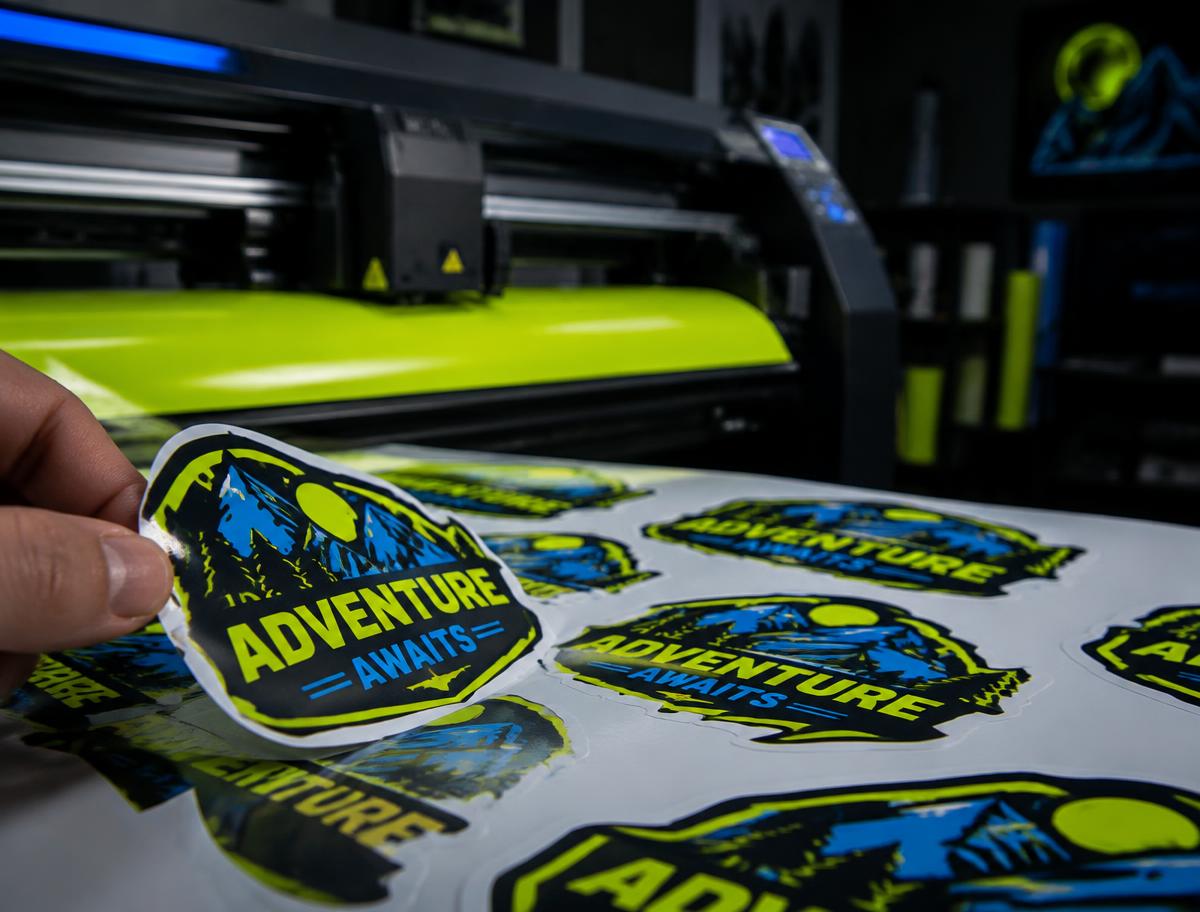 Hand peeling a die-cut sticker next to a vinyl cutting plotter
